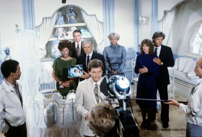 Blake Carrington (John Forsythe), con uno de sus impolutos trajes a medida, junto a Alexis (Joan Collins) muestran la foto de un bebé rodeados por el resto de la familia en una habitación de tules y molduras preparada para un bebé de alta cuna. |