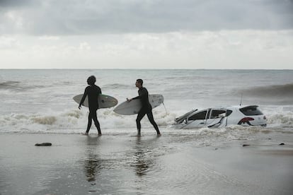 Dos surfistas pasan delante de un coche arrastro por el temporal.