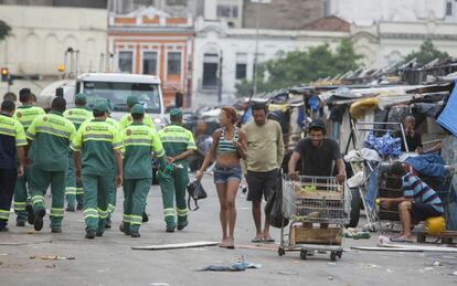 Moradores da cracolândia passam pela equipe de limpeza da Prefeitura de São Paulo. Ao fundo, mais barracos e lixo pelo chão.