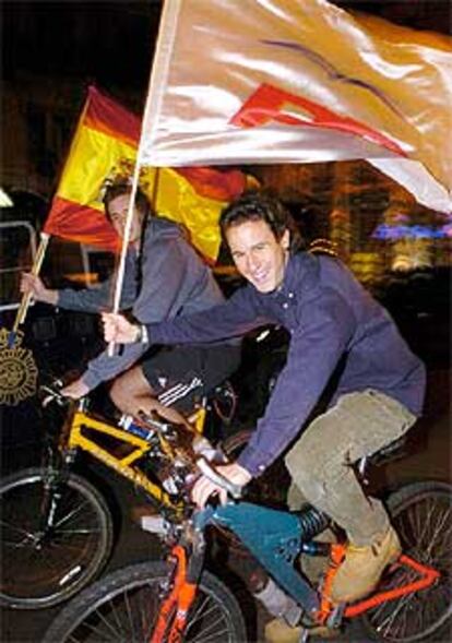 A pesar de que su partido perdió las elecciones generales, dos jóvenes se muestras contentos mientras recorren en bicicleta las inmediaciones de la calle de Génova portando una bandera de España y otra del PP.