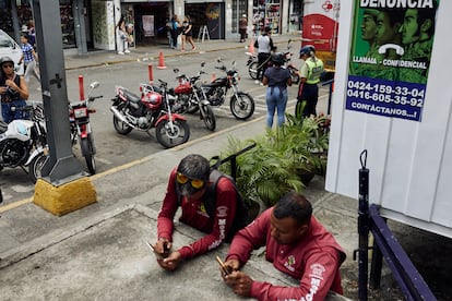 Dos hombres usan su teléfono móvil mientras una policía trabaja tras ellos, en Caracas (Venezuela), el 28 de julio de 2024.