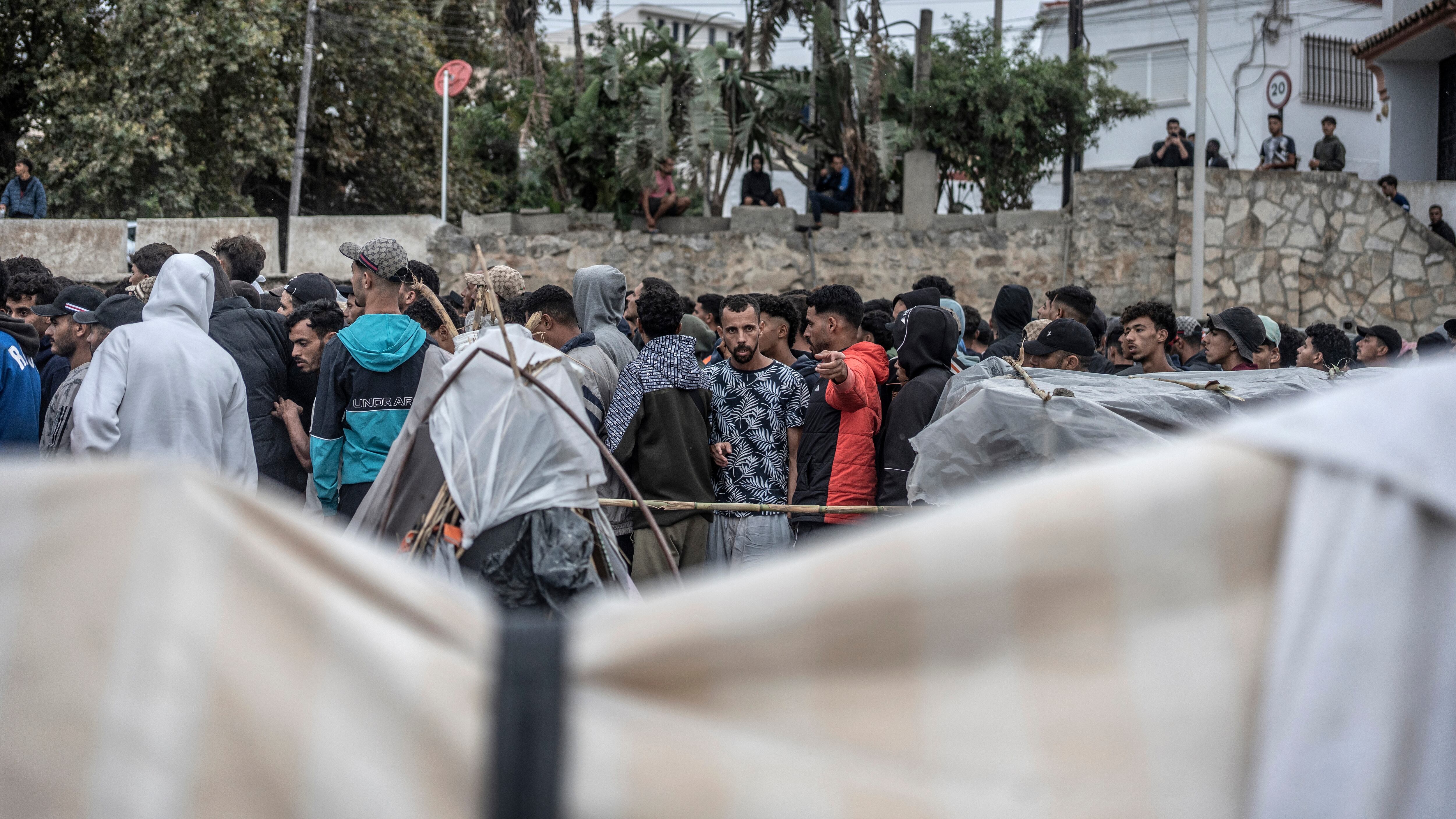 Migrantes marroquíes en su mayoría hacen cola para acceder al cordón policial que da acceso al reparto de comida organizado por Cruz Roja en la playa del Trampolín en Ceuta, este lunes.