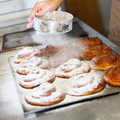 Baker hands dusting freshly baked ensaimadas with powdered sugar in bakery. Baking process of traditional Spanish pastries