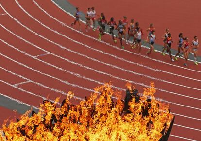 Las atletas pasan en frente de la llama olímpica en la competición de los 5.000 metros.