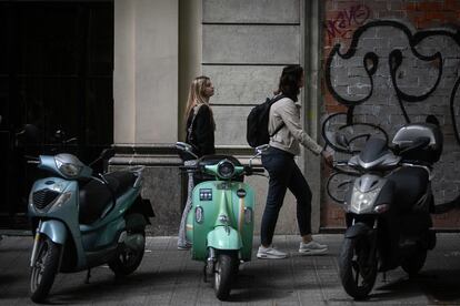 Dos jóvenes caminan por una acera estrecha de Barcelona donde hay varias motos aparcadas. 