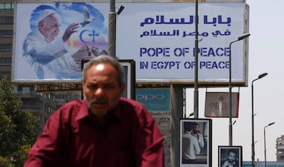 Un hombre conduce su bicicleta en el cartel de la visita del Papa a El Cairo al fondo. 