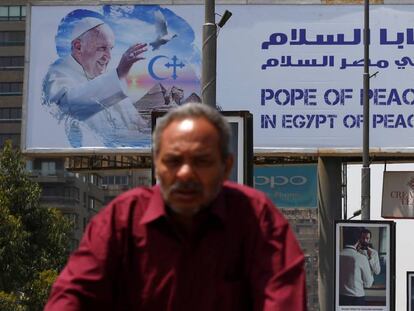 Un hombre conduce su bicicleta en el cartel de la visita del Papa a El Cairo al fondo. 