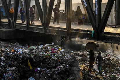 Una mujer transporta desechos sobre pilas de basura debajo de un puente peatonal en Dhaka (Bangladés)