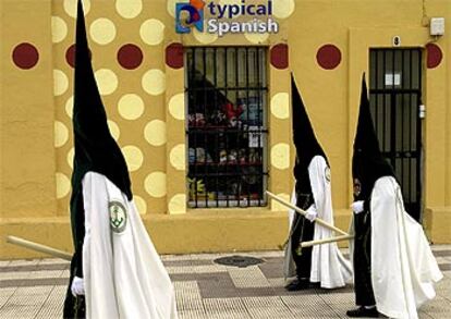 Tres nazarenos de la Hermandad de la Esperanza de Triana pasan junto a una tienda de recuerdos a primera hora de la mañana. La lluvia truncó la <i>Madrugá</i> del Viernes Santo en Sevilla, que se convirtió en atípica y trianera, ya que la Hermandad de la Esperanza de Triana fue la única de las seis cofradías que hizo su estación de penitencia en una noche que comenzó con lluvias y que terminó con claros.