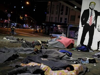 Moradores de rua descansam em frente a um grafite representando Bolsonaro e Trump.