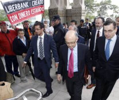 El ministro de Hacienda, Cristóbal Montoro, pasa junto a un un grupo de afectados por las preferentes que se concentraron a la entrada de la Universidad Internacional Menéndez Pelayo donde hoy se celebró el seminario " Competitividad y Marca España ".