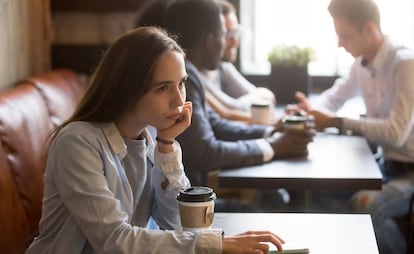 Una joven, sentada sola en un café. Detrás, un grupo de personas comparte una mesa.