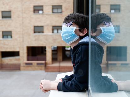 Un niño con una mascarilla se asoma a la ventana de su casa.