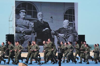 Un grupo de actores vestidos de soldado corre delante de la imagen de Winston Churchill, Franklin Roosevelt y Joseph Stalin en una pantalla gigante.