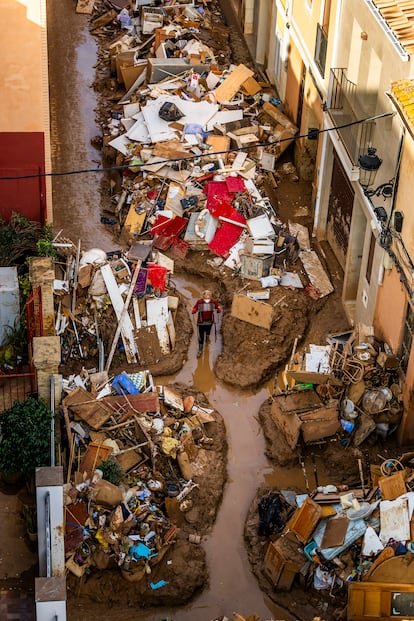 Efectos de la dana en la calle Metge Peset, en Paiporta, el 8 de noviembre de 2024. La imagen muestra la devastación provocada por la acumulación de barro, muebles y escombros tras las lluvias torrenciales. Desde una azotea se aprecia un estrecho pasillo improvisado entre los restos, utilizado como camino por los vecinos. Una mujer, cargando una silla, aporta perspectiva y escala a la escena.