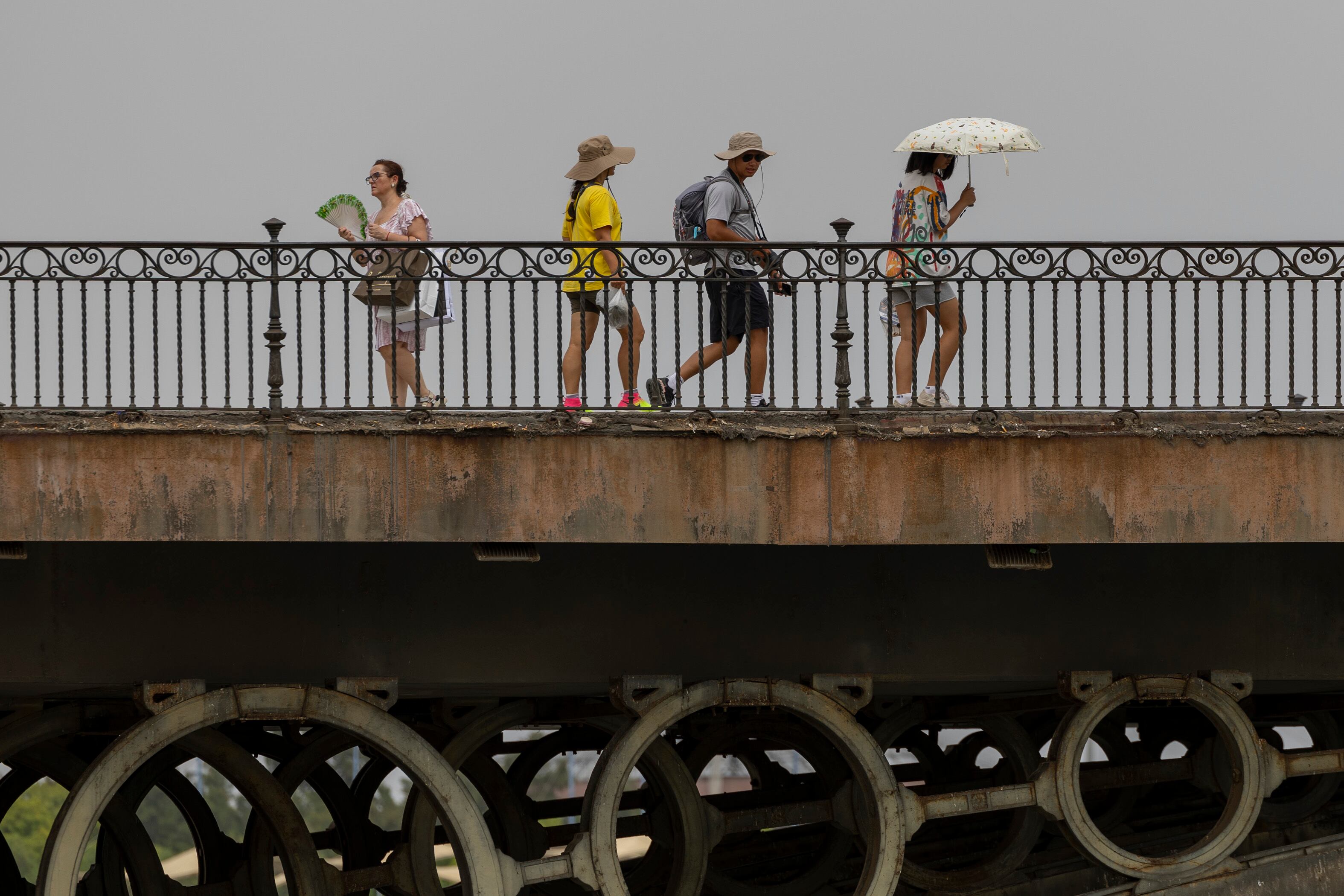 Varias personas caminan por el Puente de Triana en Sevilla este miércoles, y se protegen del calor con abanico, sombreros y paraguas.