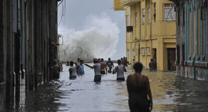 Unos cubanos caminan por una calle inundada mientras una ola rompe contra el Malecón.