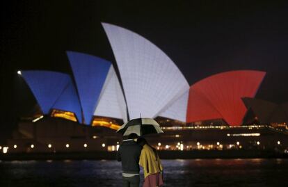 Una pareja se cubre de la lluvia en Sidney (Australia). Al fondo, la Ópera de Sidney alumbrada con los colores nacionales de Francia, en solidaridad con los atentados que han costado la vida a 127 personas en París.