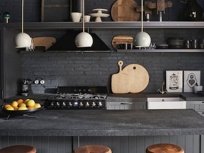 En el recibidor de la casa, sin luz natural, Beriestain apostó por ubicar la cocina para marcar un ambiente acogedor y práctico. El color gris antracita aporta más calidez a la luz artificial. Las lámparas vintage son de su tienda. La encimera es de granito negro flameado y cepillado. Los taburetes son también vintage. Las estanterías metálicas son de cocina profesional. Los cerrojos africanos, las peanas y la vajilla provienen de su concept store, y los dibujos son de Bene Rohlmann y de Peyrone.