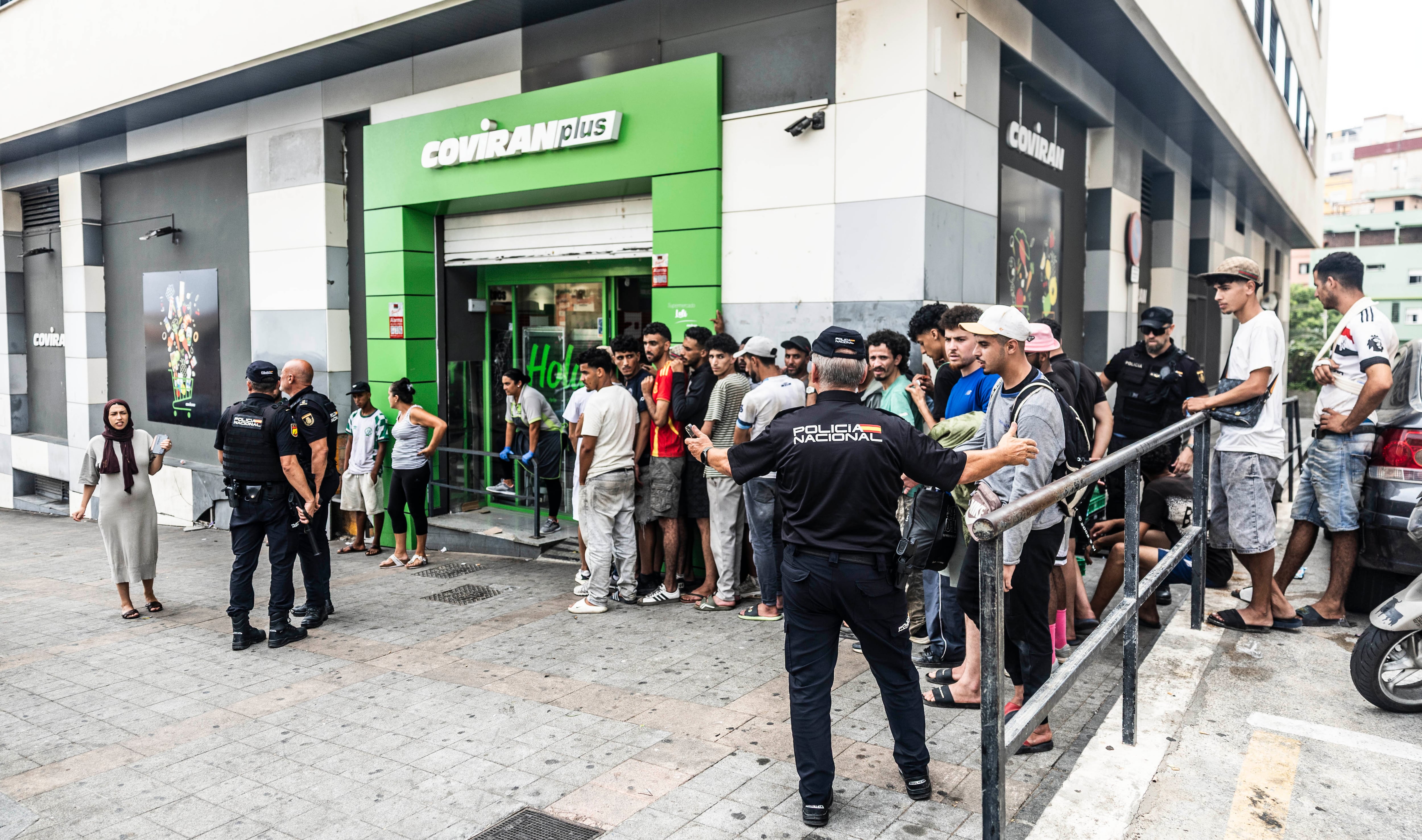 Policías nacionales preguntan a los migrantes que esperan para comprar en un supermercado si tienen euros y regulan su entrada en el supermercado, este sábado, en la Avenida Virgen de África, en Ceuta.