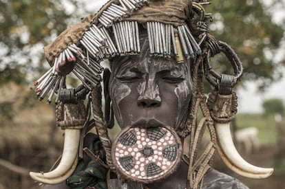 El proceso de aculturación y la emulación como modo de vida ha llevado a los mursi a adoptar tocados o vestimentas que nada tienen que ver con su tradición ni cultura. En la imagen, una mujer joven con el típico plato labial y el rostro pintado con arcilla blanca posa con un tocado compuesto por el arreo de una vaca. El contraste entre la tradición y el progreso se manifiesta claramente en los atuendos y decoraciones personales.