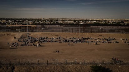 A group of migrants waits next to the border wall.