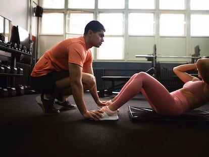 Una joven hace abdominales en un gimnasio, ayudada de su entrenador.