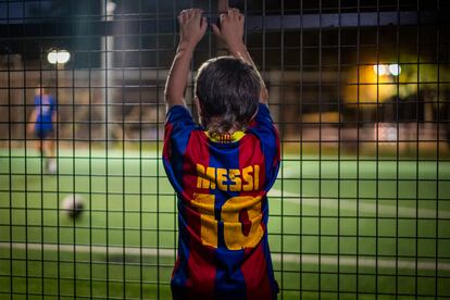 Un niño con la camiseta de Messi observa un partido en Girona a través de una valla.