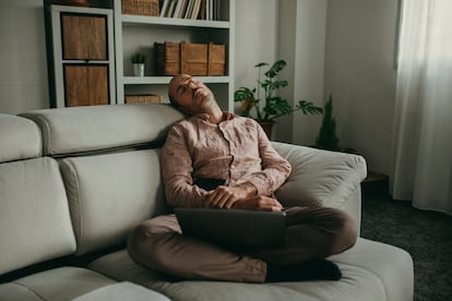 Un hombre duerme, con un portátil encima, sentado en el sofá.