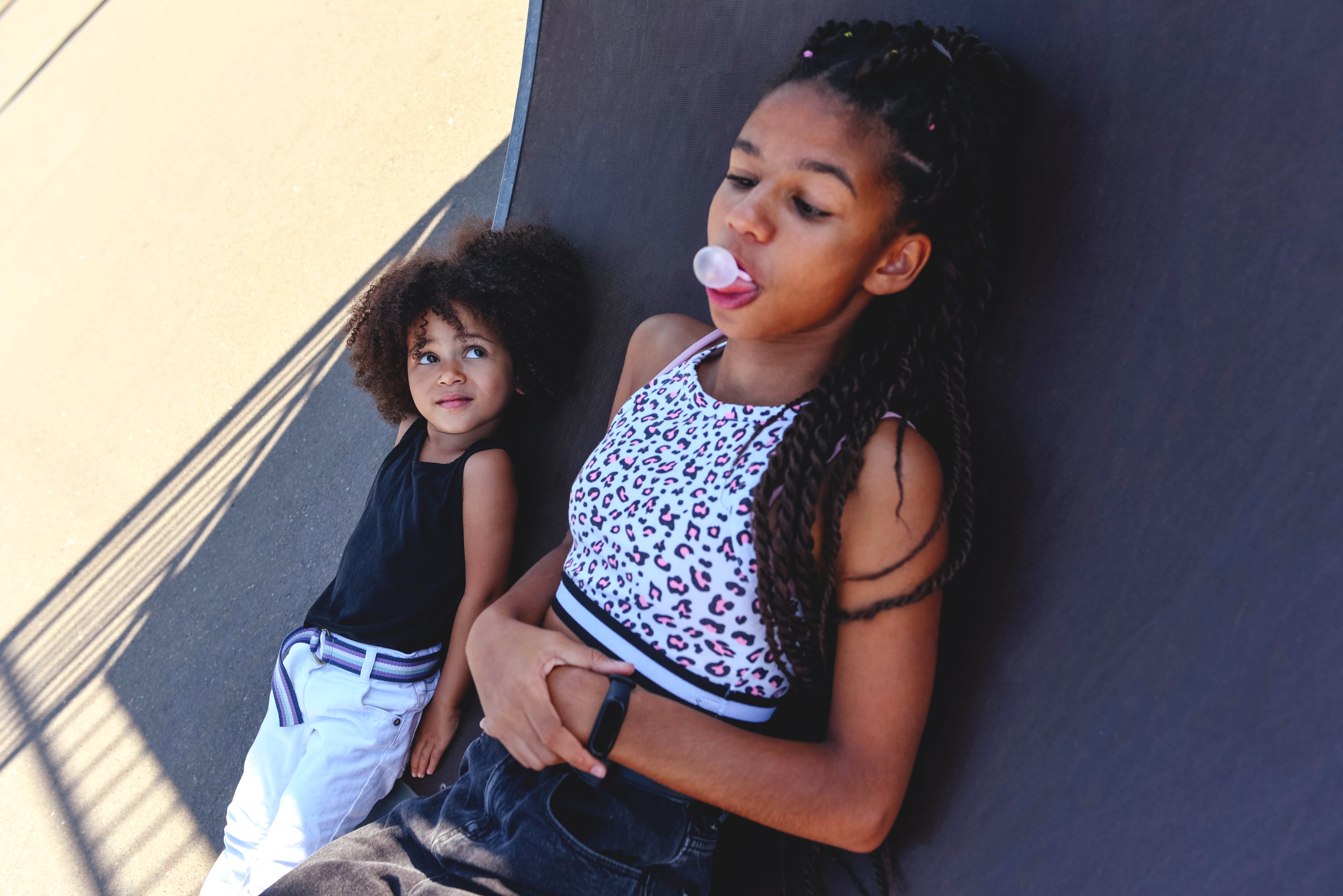 Two sisters are lying on a skateboard and chewing gum. Portraits of black girls.