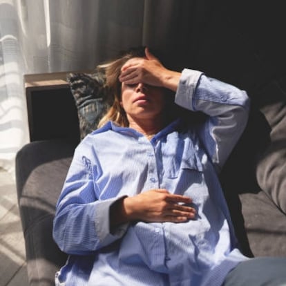 A woman lying on a couch with her hand on her forehead and eyes closed, indicating exhaustion or stress. The indoor scene is brightly lit by natural light coming through the window with curtains.
