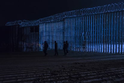 Um grupo de pessoas caminha diante do muro fronteiriço de Tijuana, na sexta-feira à noite.