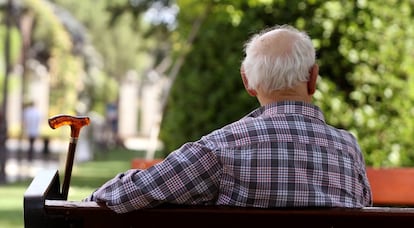 Un anciano de espaldas sentado en un banco junto a su bastón. 