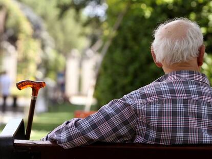 Un anciano de espaldas sentado en un banco junto a su bastón. 