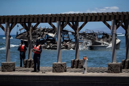 U.S. Coast Guards stand in front of the burnt carcass of a boat sinking in the port after a wildfire burned through the city up to the port in Lahaina.