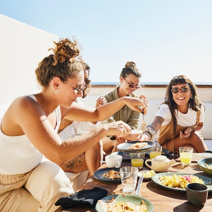 Medium wide shot of smiling female friends sharing breakfast on deck of luxury suite at tropical resort