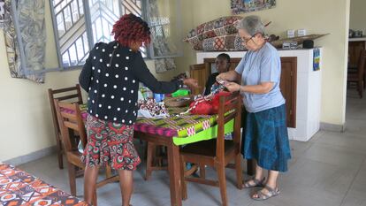 Una mujer está sentada en una mesa mientras Sor Ángela, a la derecha de la imagen, ayuda a otra mujer con unas telas