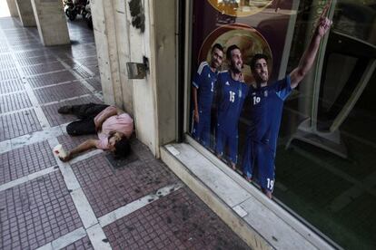 Un hombre pide dinero en una calle de Atenas frente a un cartel que promociona a la selecci&oacute;n de Grecia.