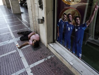 Un hombre pide dinero en una calle de Atenas frente a un cartel que promociona a la selecci&oacute;n de Grecia.