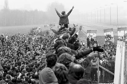 Entre una multitud encaramada al muro de Berlín, un joven en pie levanta los brazos y hace el signo de la victoria con ambas manos. A la izquierda se observa la plaza de Postdamer repleta de gente.