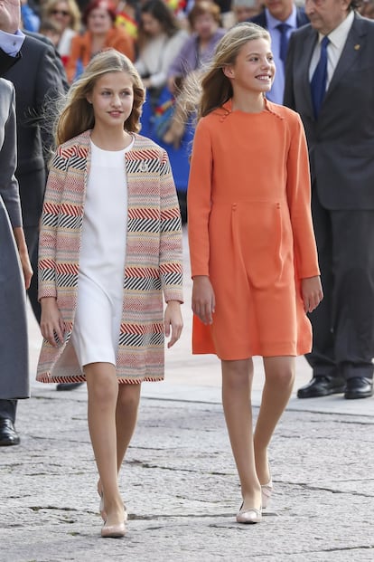 La princesa Leonor, con el pelo suelto, ha vestido un sencillo 'look' compuesto por un vestido blanco sobre el cual se ha puesto un abrigo de rayas en tonos rosas y azules. Su hermana, la infanta Sofía, ha lucido un vestido naranja vivo de manga larga.