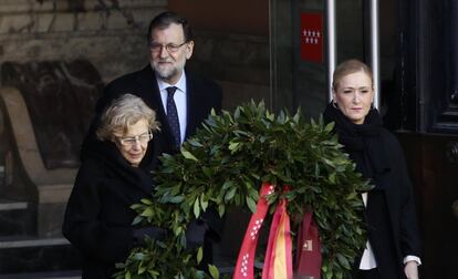 La presidenta de la Comunidad de Madrid, Cristina Cifuentes (i), y la alcaldesa de Madrid, Manuela Carmena, llevan una corona de flores en el acto de homenaje en la Puerta del Sol a las víctimas del 11-M. Detrás, el presidente del Gobierno en funciones, Mariano Rajoy, el 11 de marzo de 2016.