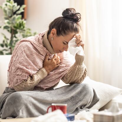 Very sick young woman sitting on a couch holding her forearm and handkerchief at her nose fighting fever.