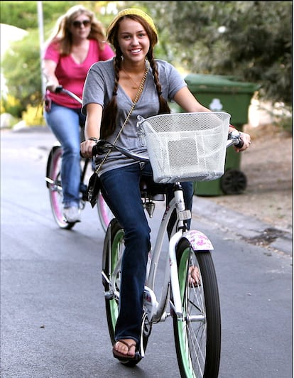 Miley y su bici. La hemos visto madurar sobre dos ruedas y aquí muestra un clásico de sus estilismos tempranos: dos trenzas, gorrito, camiseta básica de algodón y un buen par de jeans.