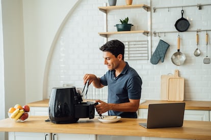 Un hombre usa una freidora de aire en su cocina, mientras consulta la receta en su portátil.