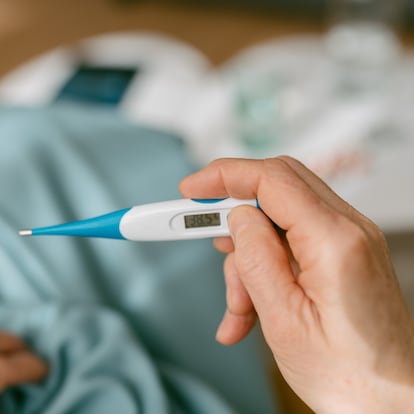 Over-the-shoulder view of an unrecognizable woman looking at the thermometer because she is sick while sitting on the sofa covered with a blanket.