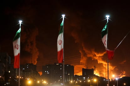 Banderas iraníes ondean mientras el fuego y el humo se elevan desde el depósito petrolero de Sharan, tras un ataque israelí este domingo.