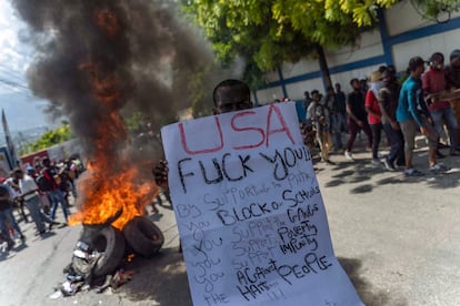 Además de la escalada de los precios, el regreso de la enfermedad del cólera a Haití, luego de su erradicación hace 10 años, ha complicado aún más la situación en la nación caribeña. En la imagen, un manifestante sostiene un cartel en contra de la propuesta de intervención militar extranjera, junto a una pila de neumáticos incendiados.