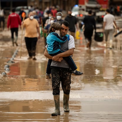 En la imagen, un hombre carga con un niño mientras cruza un puente lleno de agua bajo el barranco del Poyo en Valencia.