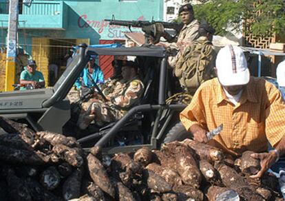 Un hombre vende yuca en un mercado de Santo Domingo mientras una patrulla militar vigila la zona.